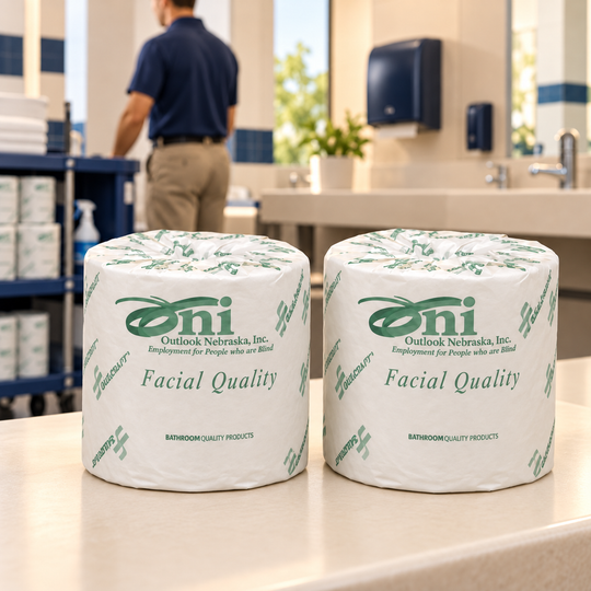 Two individually wrapped toilet tissue rolls standing upright side by side on a restroom counter, with a male facilities worker and supply cart in the background.