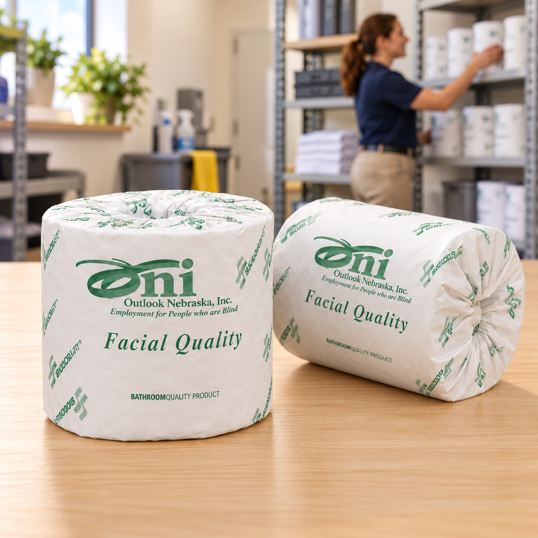 Two individually wrapped toilet tissue rolls on a wooden supply-room counter, one upright and one on its side, with a woman stocking shelves in the background.
